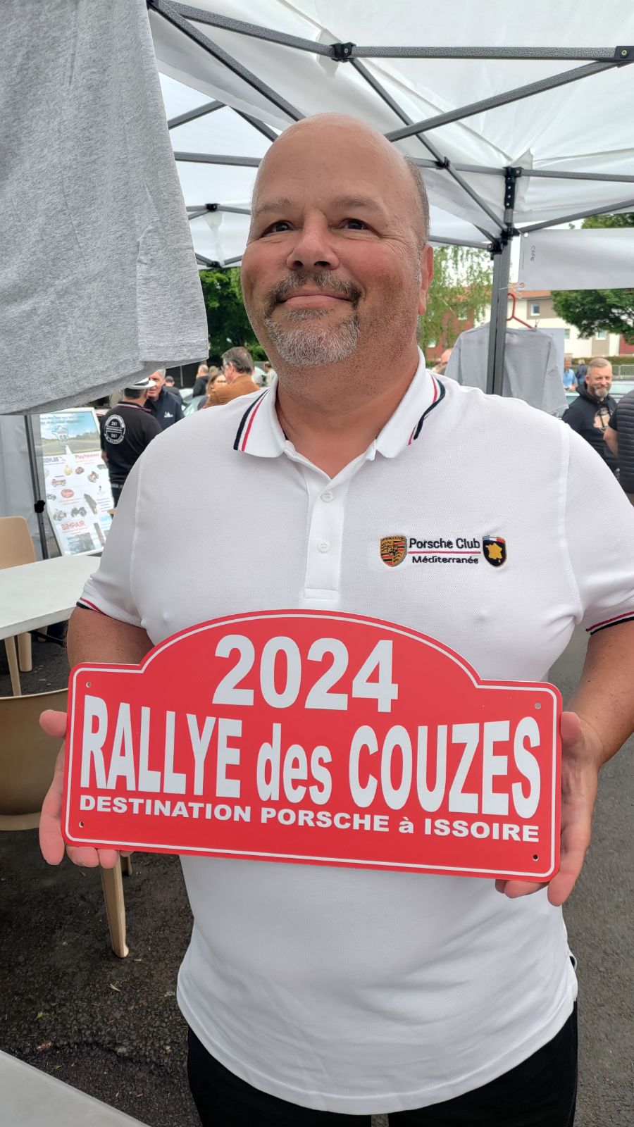 Une personne portant un polo blanc tient une pancarte sur laquelle est écrit « 2024 RALLYE des COUZES DESTINATION PORSCHE À ISSOIRE » sous un auvent lors de l'événement en plein air de 2023.