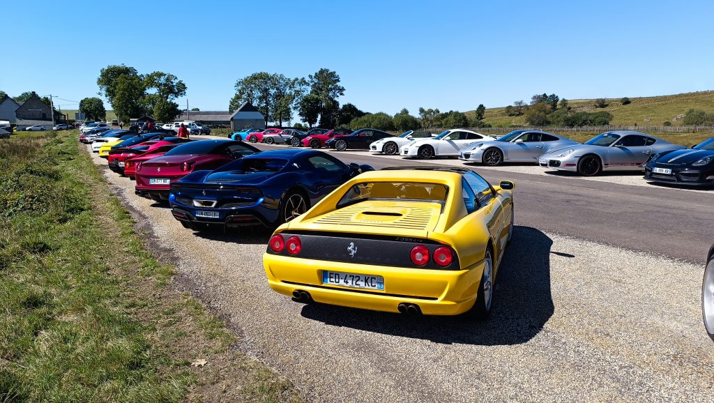 Une rangée de voitures de sport, dont une Ferrari jaune, s'aligne sur un terrain découvert sous le soleil lors d'un supercarstour, avec des arbres et des collines herbeuses en arrière-plan. porsche auvergne