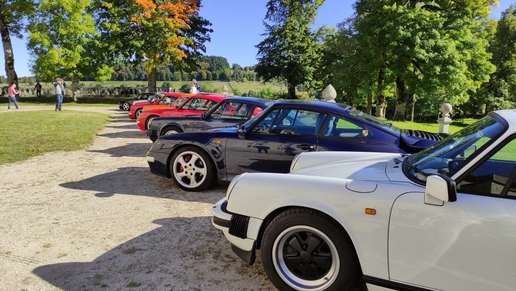 Une rangée de voitures Porsche classiques est garée dans une allée ombragée, capturant l'atmosphère animée d'une garden party avec des gens qui se mélangent et des arbres verts en arrière-plan. porsche auvergne