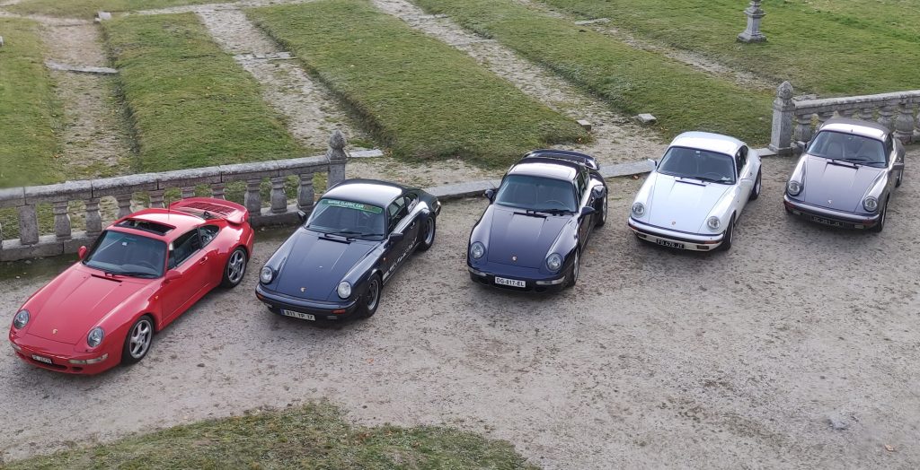 Cinq Porsche 911 de différentes couleurs sont garées en rang sur un terrain en gravier à côté d'une clôture en pierre, comme si elles étaient prêtes pour une garden-party exclusive avec de l'herbe en arrière-plan. porsche auvergne