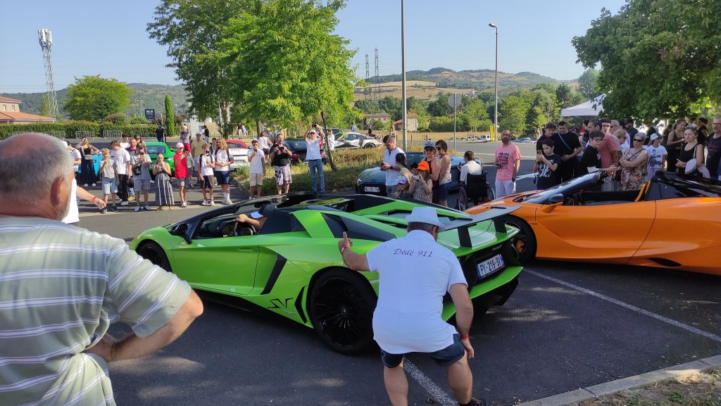 Un rassemblement se forme sur un parking, une foule se forme autour d'une voiture de sport verte et d'une voiture de sport orange ; un homme pose pour une photo devant la voiture verte. porsche auvergne